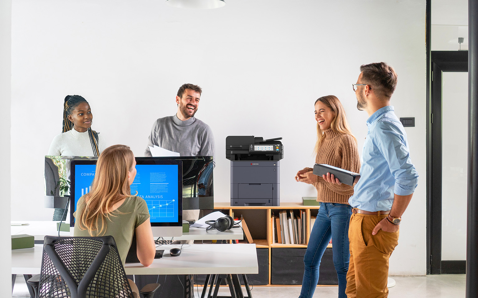 Office space with a number of employees working on different devices, such as laptops and tablets, with an MFP in the center serving as a hub for printing and document management